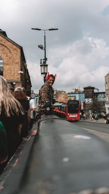 A bustling outdoor scene at Camden Market featuring multiple green and beige umbrellas with 'Camden Market' printed on their edges, shading tables and walkways filled with numerous pedestrians. In the
